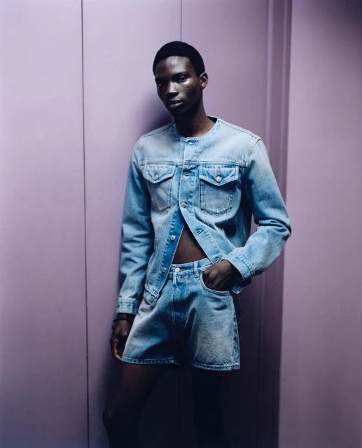 Man poses in light denim button-down and shorts against a pale purple background in a studio with soft lighting, fashion portrait.