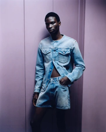 Man poses in light denim button-down and shorts against a pale purple background in a studio with soft lighting, fashion portrait.
