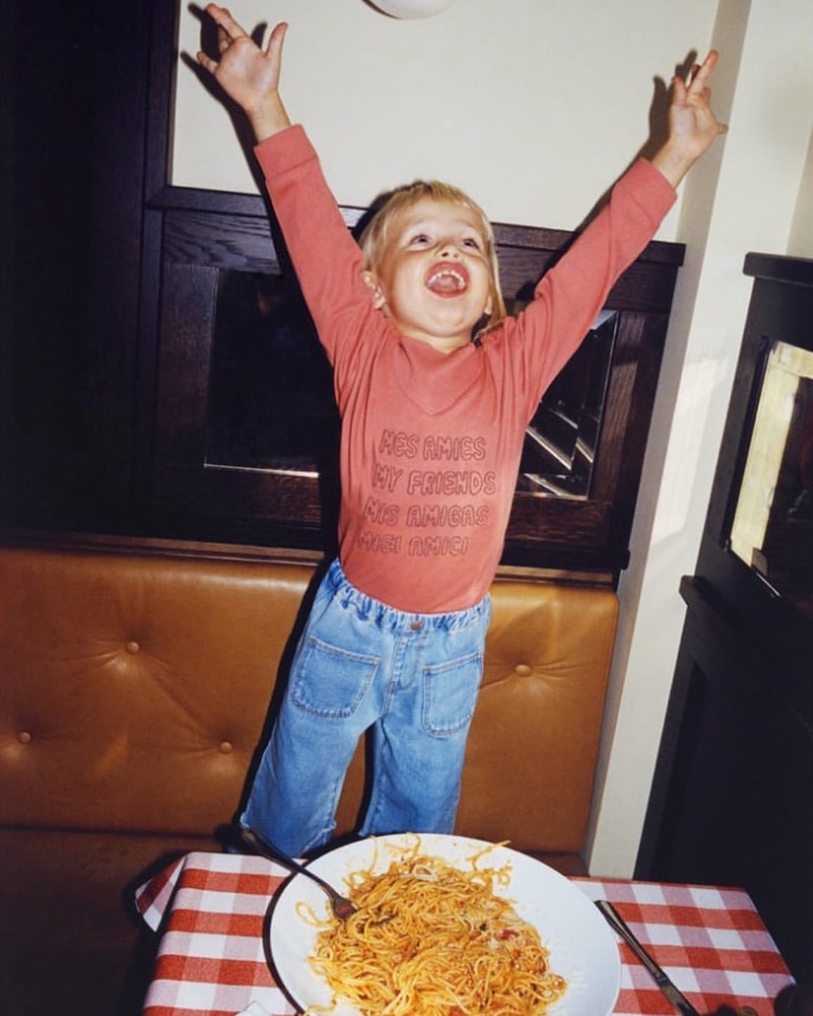 Happy child with arms raised stands near table with spaghetti and checkered tablecloth in interior setting with light streaming in.