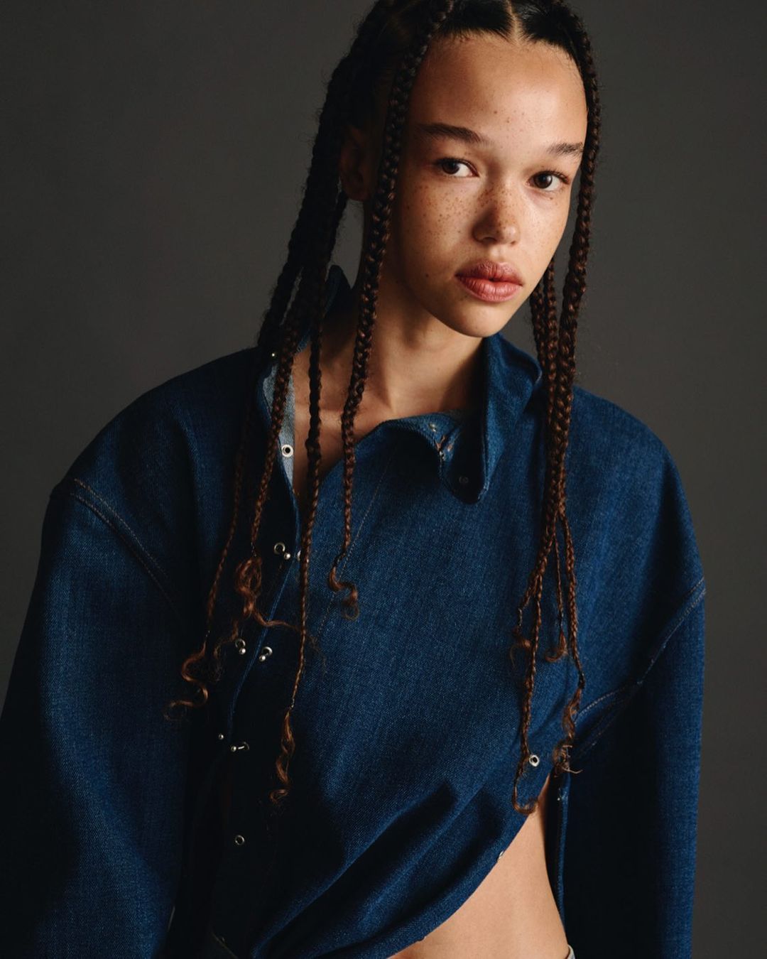 Close-up studio shot of a biracial adult woman with freckles and box braids, wearing an unbuttoned and draped denim shirt.