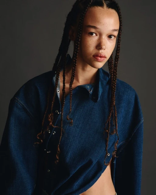 Close-up studio shot of a biracial adult woman with freckles and box braids, wearing an unbuttoned and draped denim shirt.