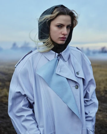 Model wearing a light blue trench coat and dark hood against a hazy outdoor field background by Luca Campri