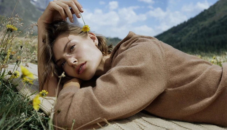 Woman in brown knit sweater lying in a meadow holding a small yellow flower near her hair, photographed by Luca Campri.