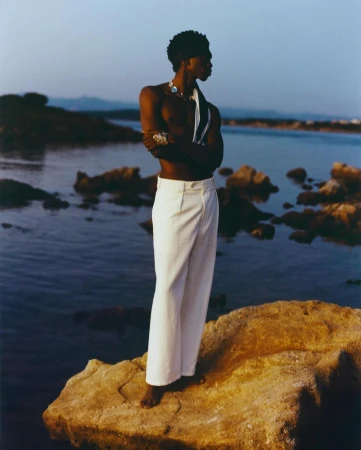 A shirtless black man wearing white trousers standing on a rocky coast at dusk by the water, photographed by Luca Campri.