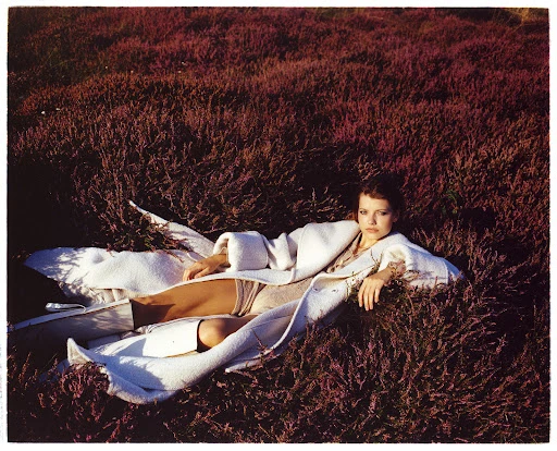 Woman in white coat lying in a field of purple heather, fashion editorial style, posing with hand near her face.