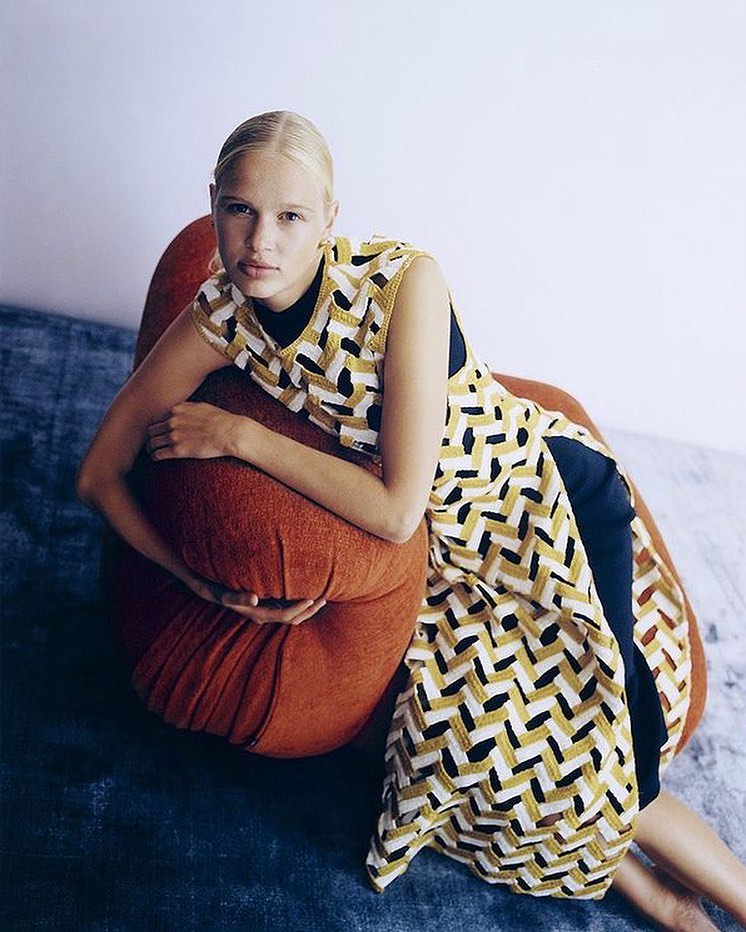 Woman in a geometric-patterned dress reclines on an orange cushion with a blue rug and white wall in a brightly-lit studio.