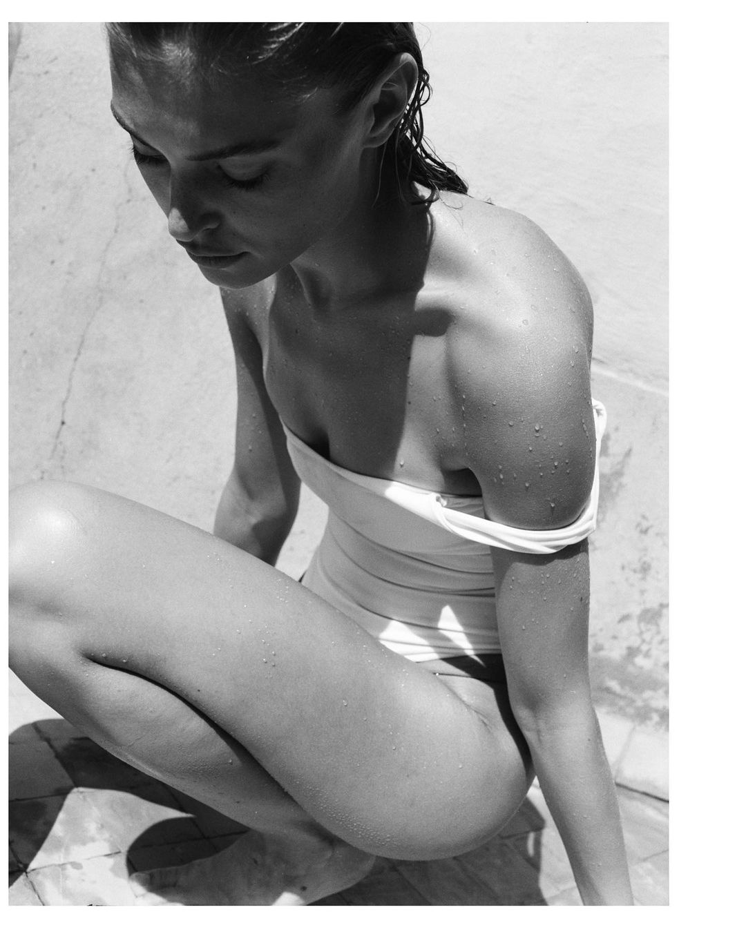 Black and white portrait of a woman in a swimsuit, crouching on a tiled surface, with wet hair and water droplets on her skin.