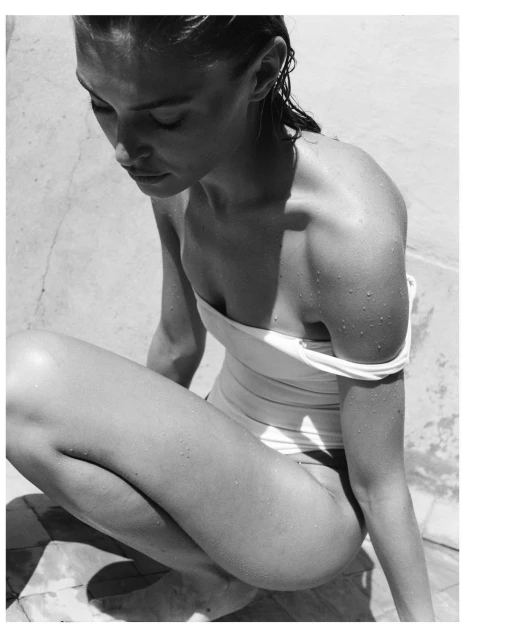 Black and white portrait of a woman in a swimsuit, crouching on a tiled surface, with wet hair and water droplets on her skin.