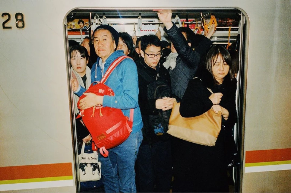 Crowded train car with diverse Asian passengers, including a man in a blue hoodie with a red backpack, creates a sense of urban density.