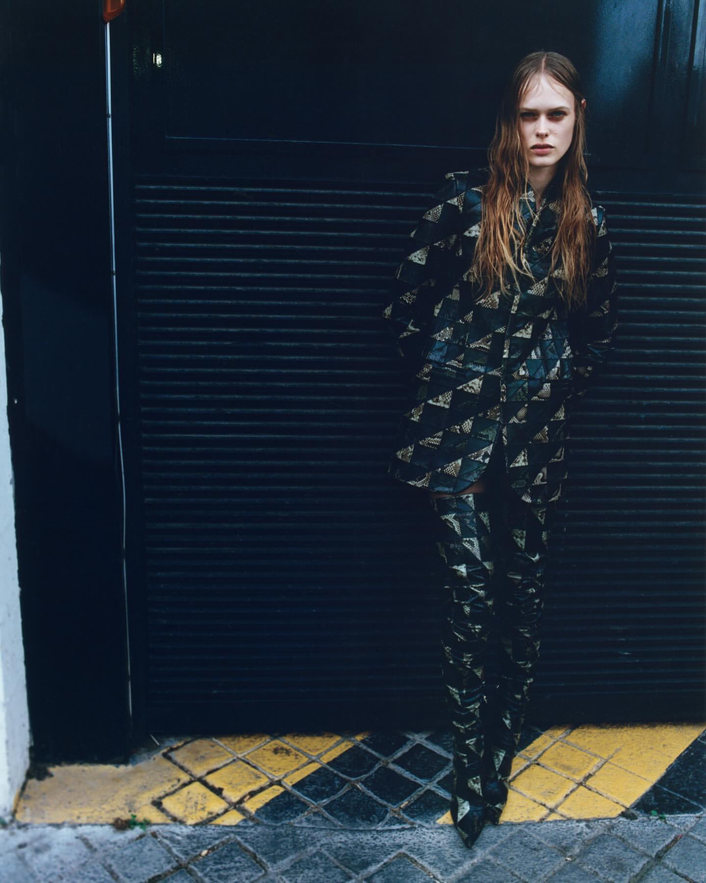 A slender woman poses in a patterned black and gold jacket and pants. She stands against a black industrial door with a wet hair look.