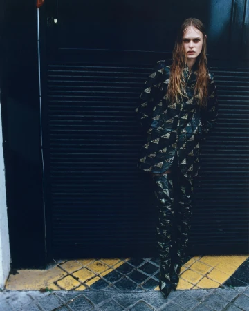 A slender woman poses in a patterned black and gold jacket and pants. She stands against a black industrial door with a wet hair look.