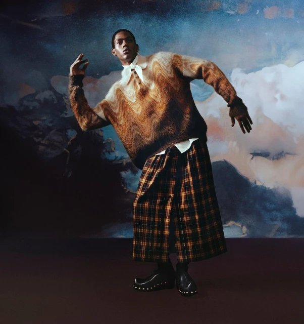 Man in textured brown sweater and plaid kilt poses dynamically against a painterly backdrop with muted, dreamlike tones in a studio setting.