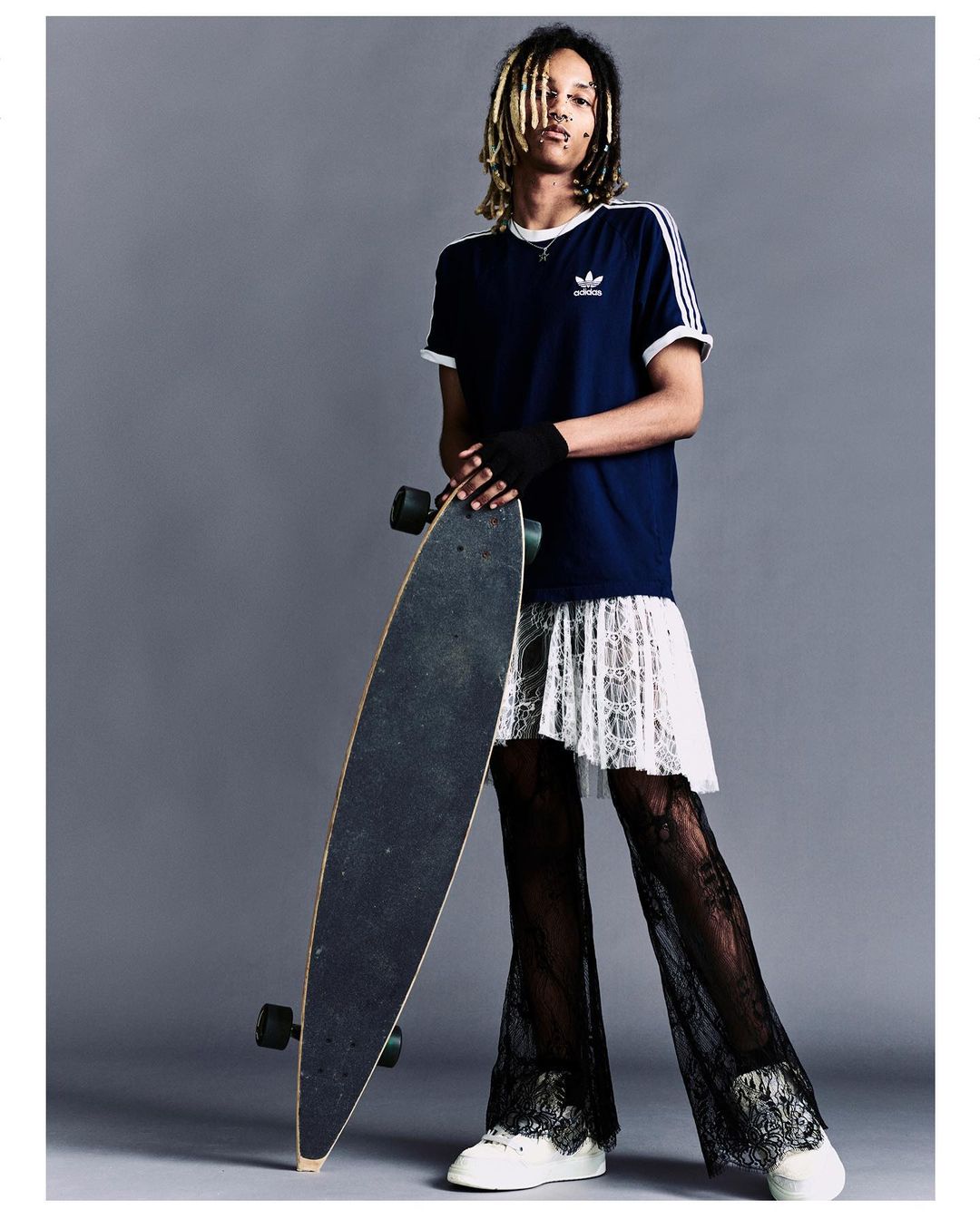 A person with dreadlocks and a black t-shirt poses holding a skateboard, wearing a white lace skirt and black lace pants in a studio.