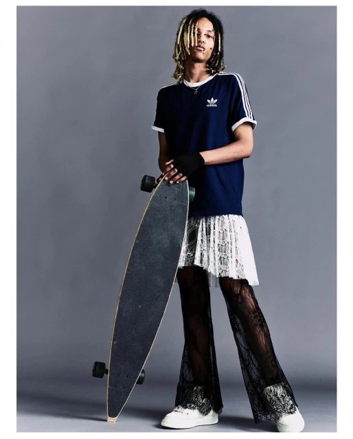 A person with dreadlocks and a black t-shirt poses holding a skateboard, wearing a white lace skirt and black lace pants in a studio.