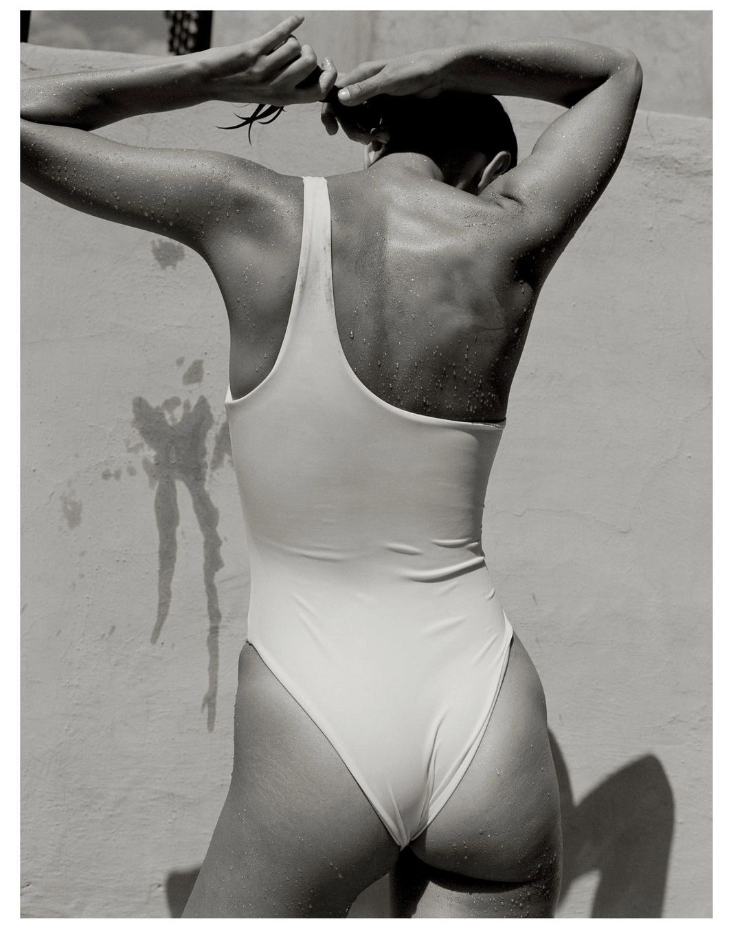 Back view of a woman in a one-piece swimsuit, with her hands raised tying her hair against a textured off-white wall, in black and white.