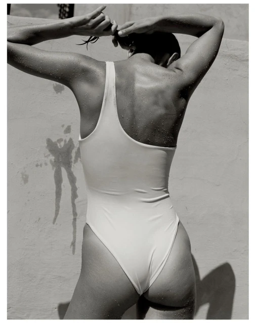Back view of a woman in a one-piece swimsuit, with her hands raised tying her hair against a textured off-white wall, in black and white.