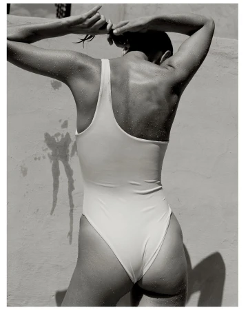 Back view of a woman in a one-piece swimsuit, with her hands raised tying her hair against a textured off-white wall, in black and white.