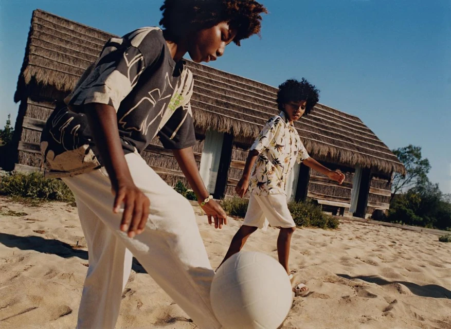 Two children, outdoors on a sandy surface, play with a ball in front of a small hut with a thatched roof on a sunny day.