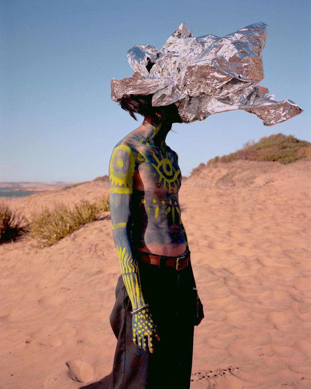 A person with yellow body paint and wearing a crumpled silver foil hat stands in a sandy, grassy landscape beneath a clear blue sky.