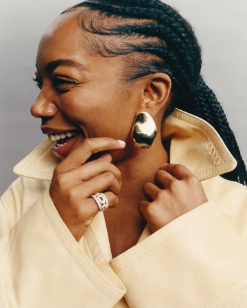 Portrait of a laughing black woman wearing large gold earrings, a diamond ring, and a cream collar jacket.