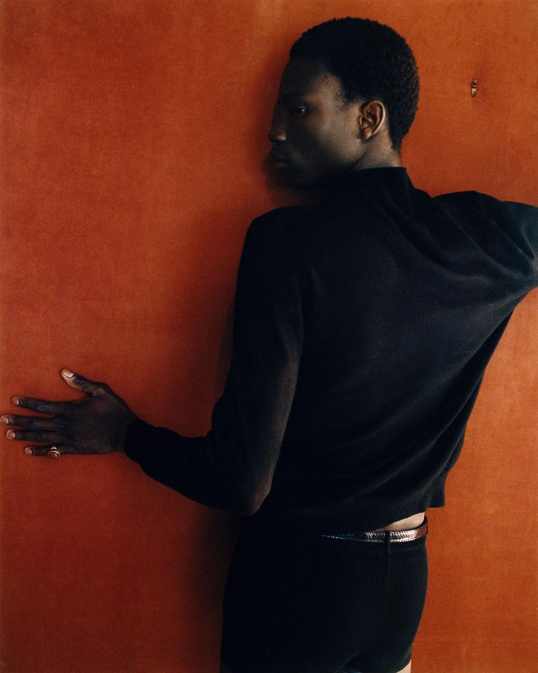An androgynous person with dark complexion poses against a textured orange background wearing a black sweater and shorts.