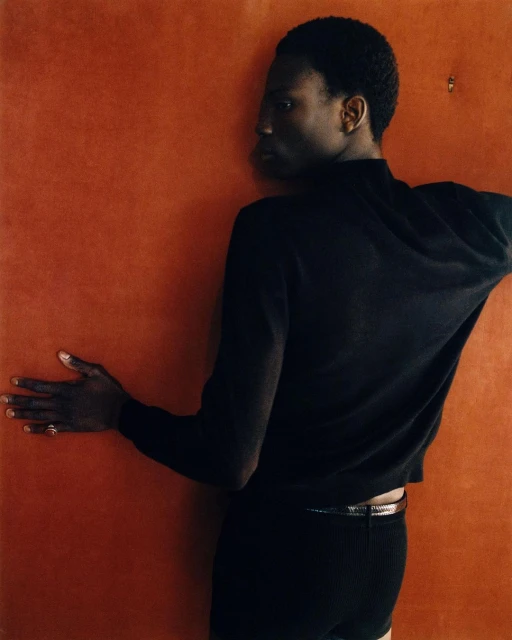 An androgynous person with dark complexion poses against a textured orange background wearing a black sweater and shorts.