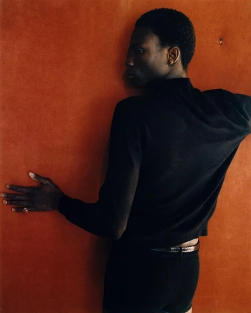 An androgynous person with dark complexion poses against a textured orange background wearing a black sweater and shorts.