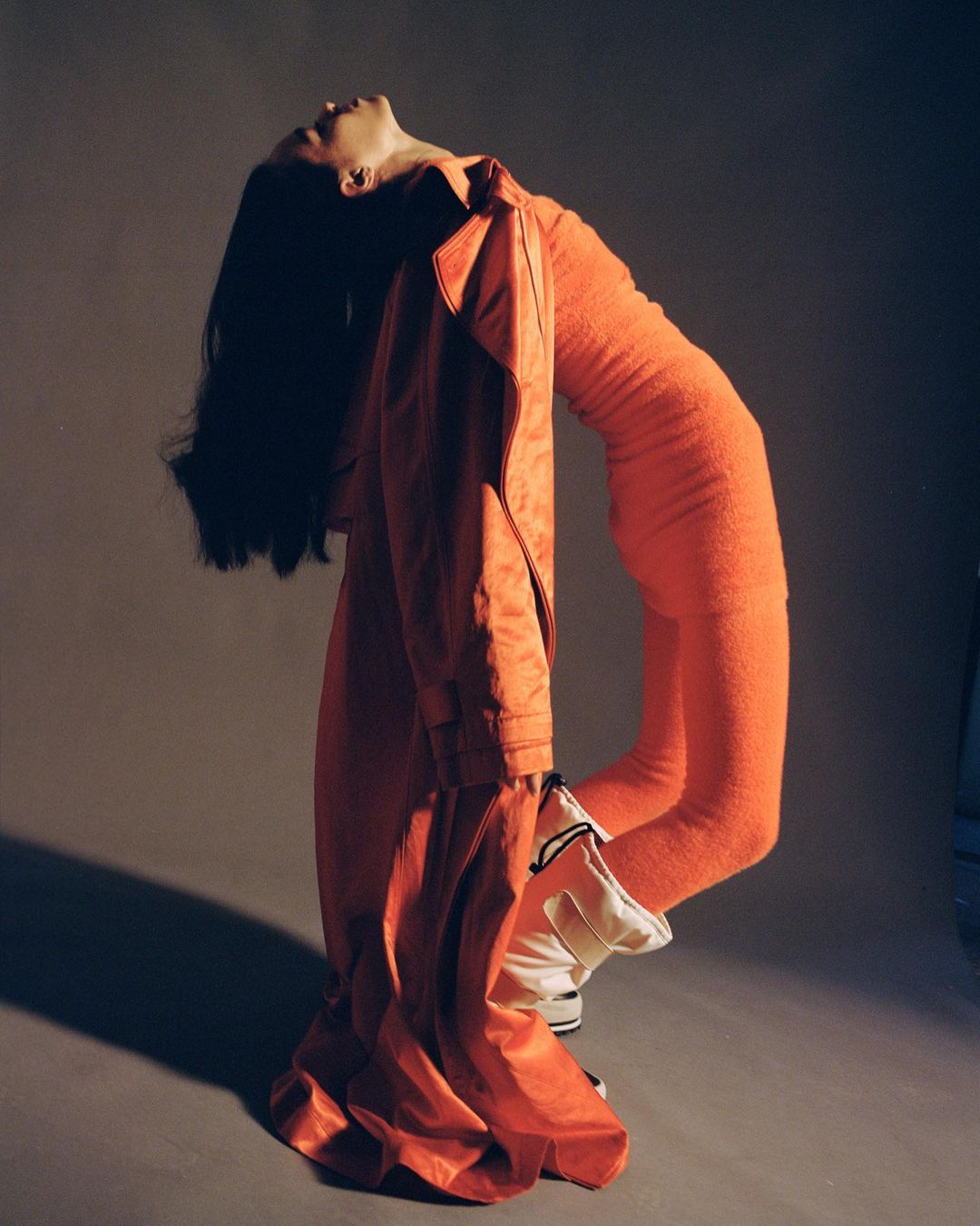 Fashion shot of a slender woman in an orange outfit arched back dramatically, with a draped orange coat in a studio setting.