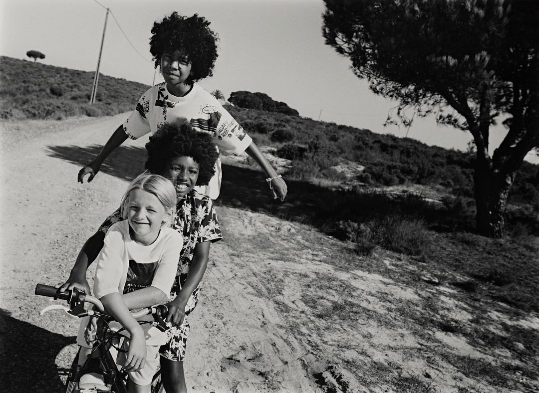 Three children pose on a bicycle on a dirt road with a grassy, tree-covered hillside in the background, shot in black and white.