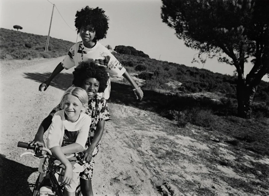 Three children pose on a bicycle on a dirt road with a grassy, tree-covered hillside in the background, shot in black and white.