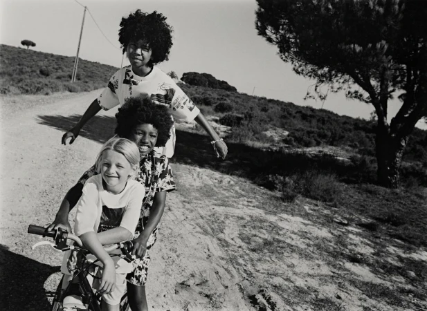 Three children pose on a bicycle on a dirt road with a grassy, tree-covered hillside in the background, shot in black and white.