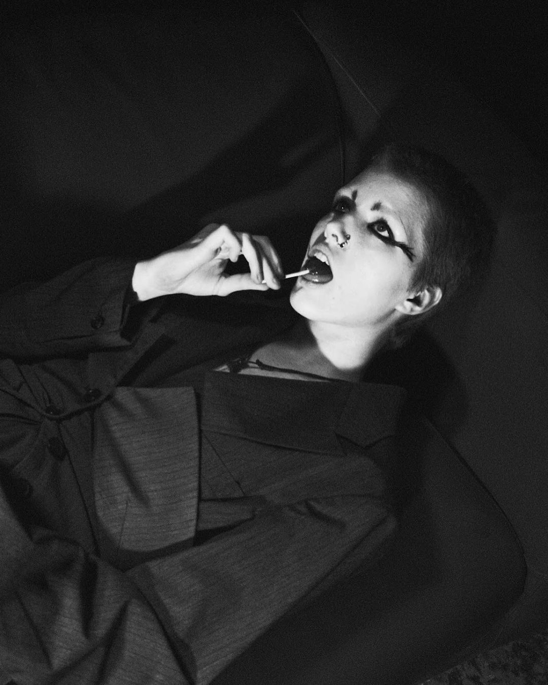 Monochromatic portrait of a young adult with dark makeup and short hair, wearing a suit and enjoying a lollipop while lying down.