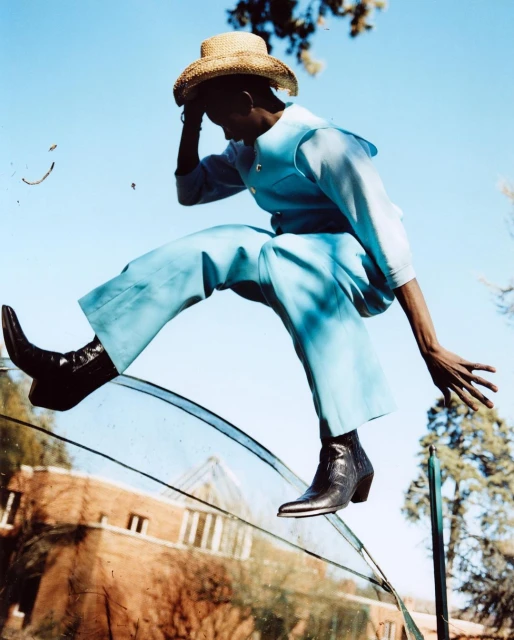 Person in blue outfit and hat jumping over a glass surface with building and trees in background on a sunny day.