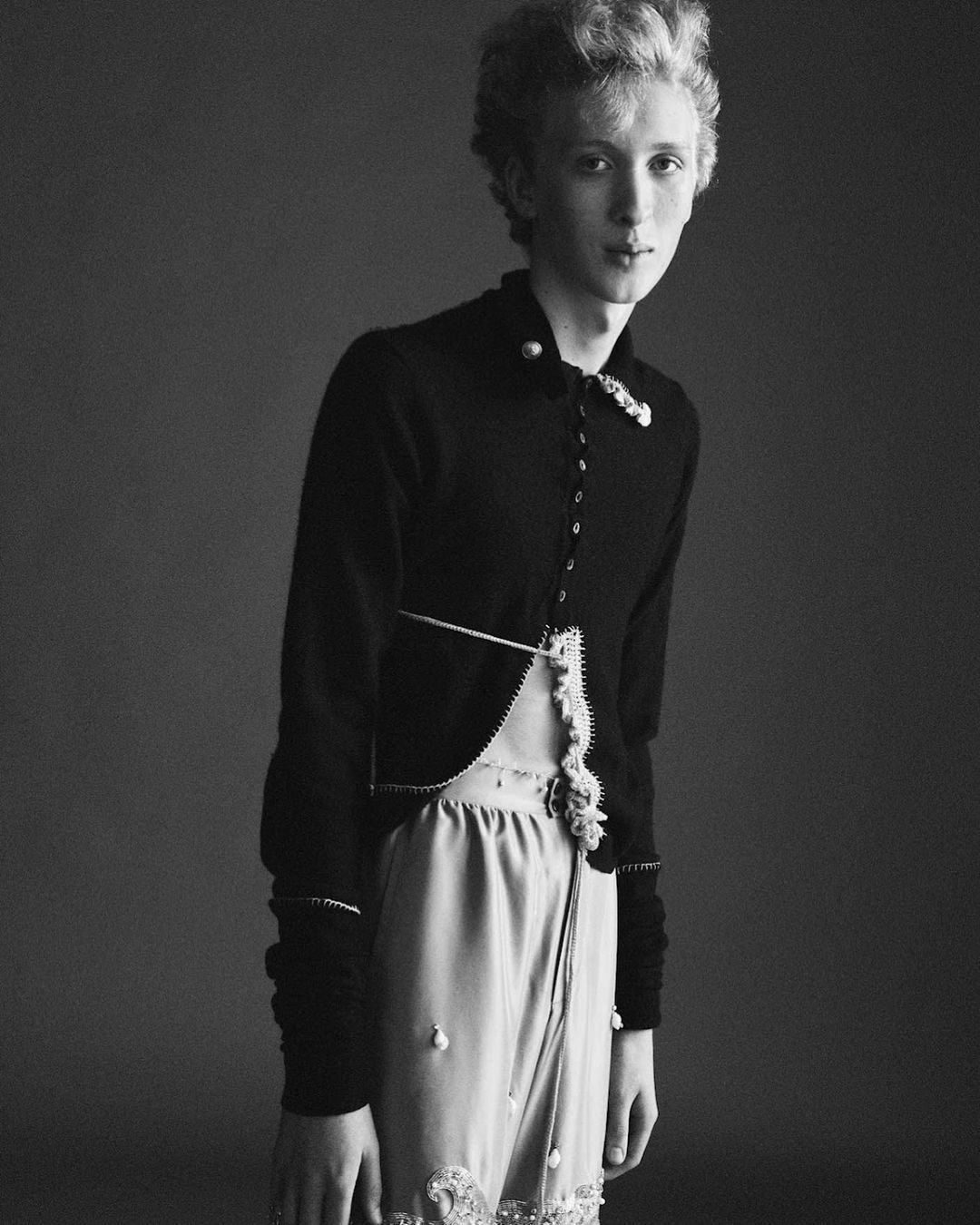 Monochrome portrait of a young adult with curly blonde hair wearing an ornate black jacket and light pink skirt in a studio.