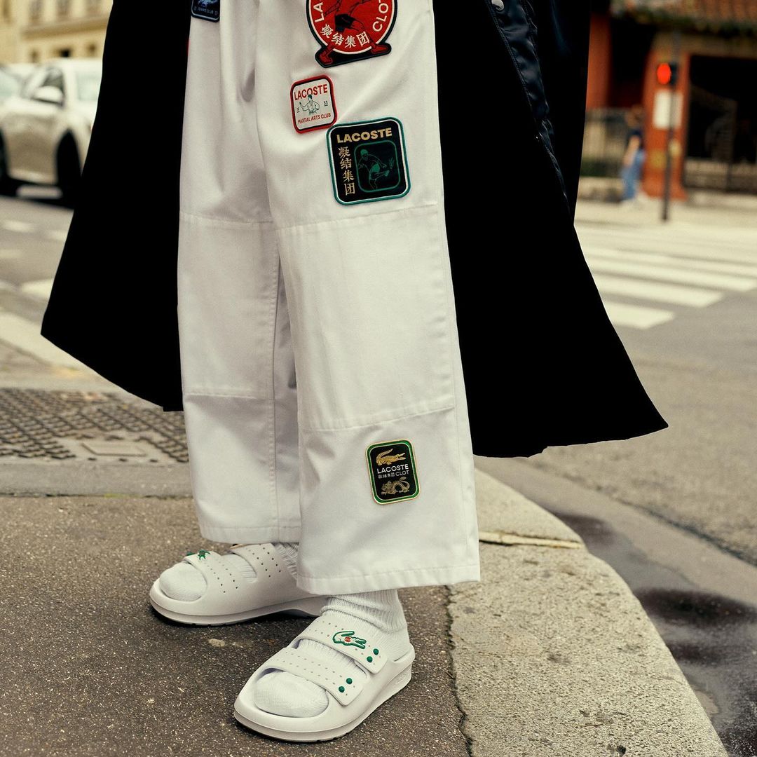 A person is wearing white pants and sandals with Lacoste patches on an outdoor city street, with a black coat draped over the shoulders.