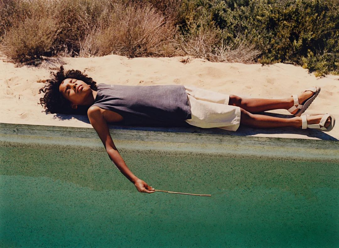 A young person with curly dark hair lying on a concrete ledge beside green water, wearing a dark tank top and light shorts.