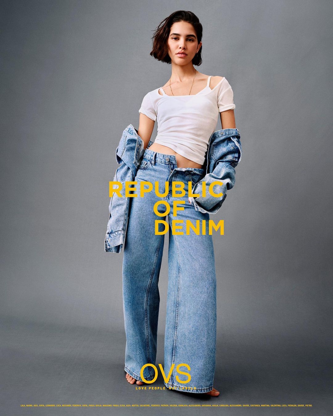 A woman models denim wide-leg jeans and a white tee shirt with a denim jacket hanging off her shoulders in a fashion shot.