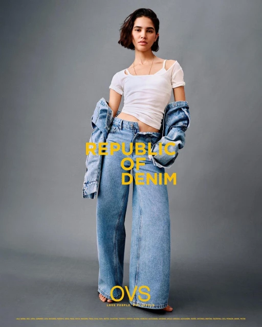 A woman models denim wide-leg jeans and a white tee shirt with a denim jacket hanging off her shoulders in a fashion shot.