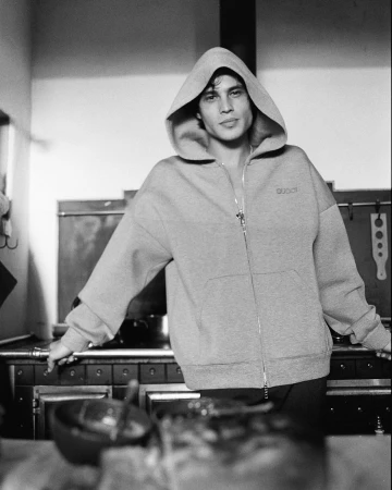 Black and white portrait of a man with dark hair wearing a hooded grey zip-up sweatshirt, standing in a kitchen, leaning on a counter.