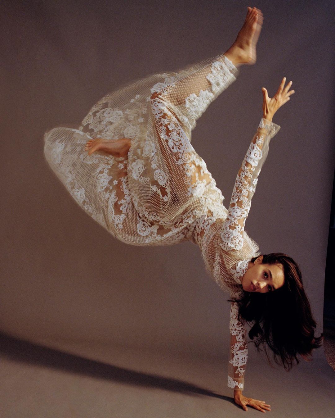 Dancer in a sheer white lace jumpsuit captured mid-air in a dynamic inversion pose against a grey seamless studio backdrop.