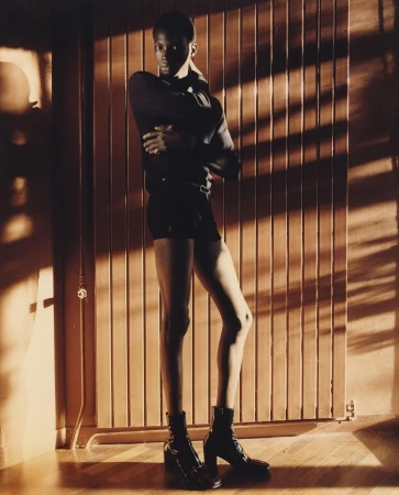 Portrait of a Black man in black clothing and heeled boots posing against a slatted wooden wall with dramatic shadows by Luca Campri.