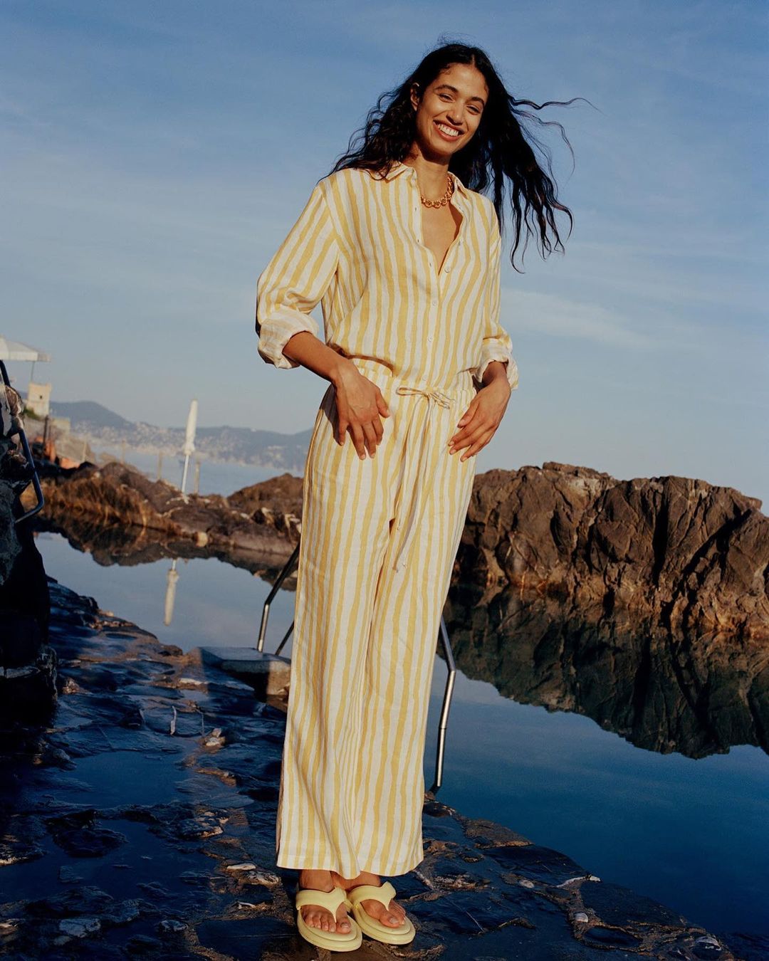 Smiling woman in a striped jumpsuit stands on rocks by the water on a sunny day, with a coastal city visible in the distance.