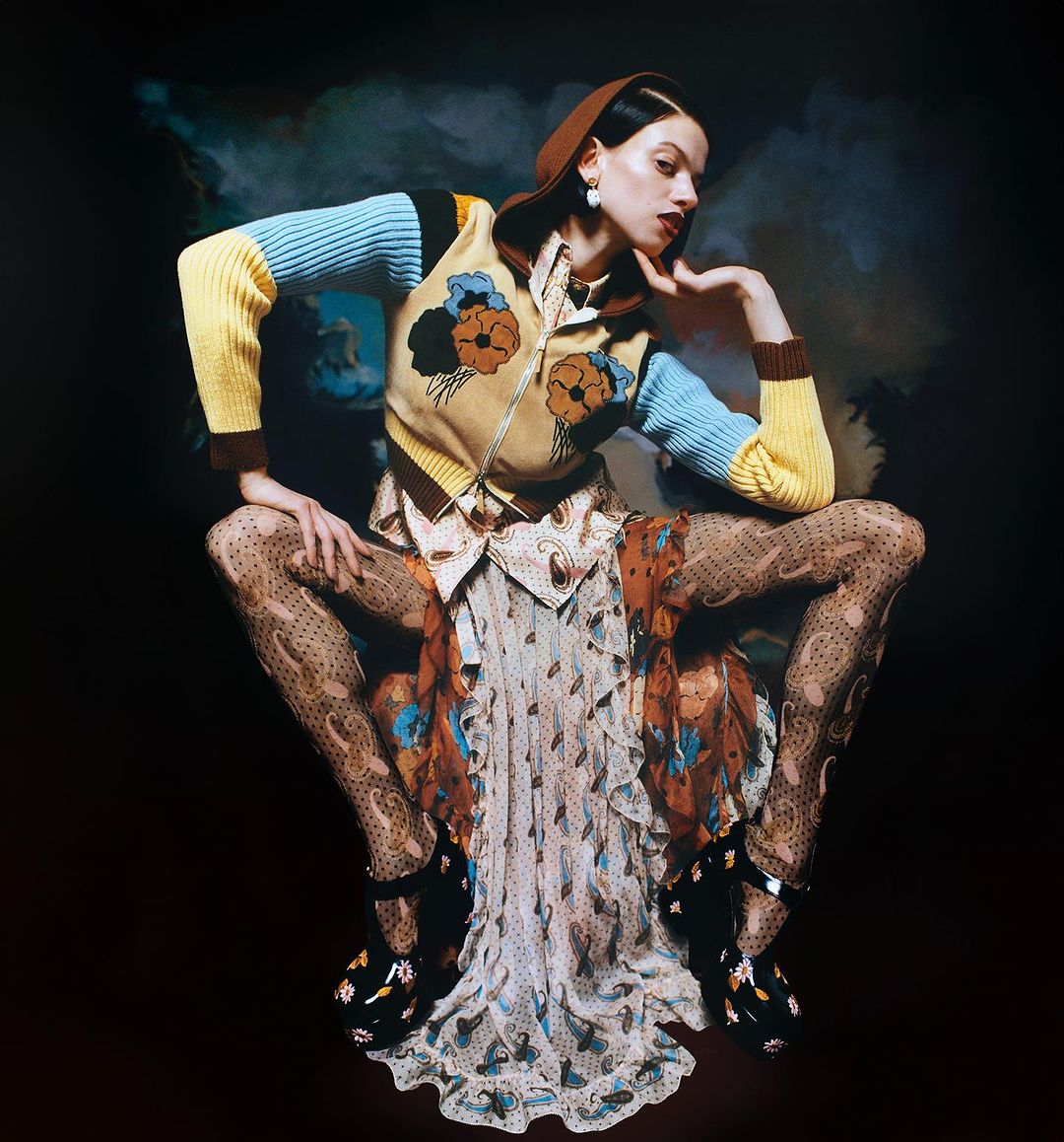 A stylish woman in patterned tights, dress, and a sweater poses against a dark backdrop with soft, cloudy accents. Beauty and fashion.
