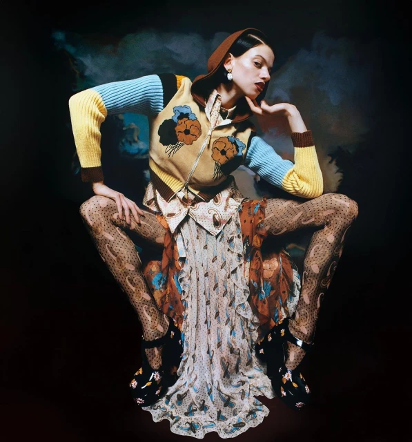 A stylish woman in patterned tights, dress, and a sweater poses against a dark backdrop with soft, cloudy accents. Beauty and fashion.
