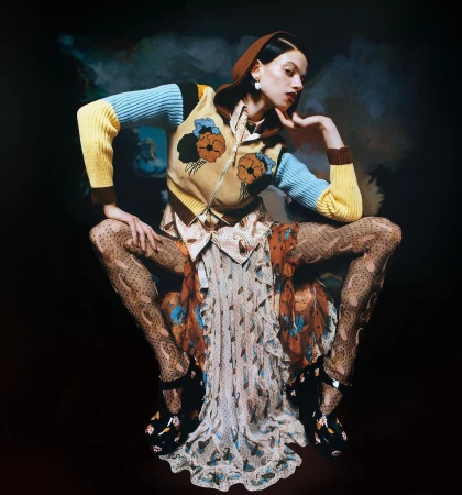 A stylish woman in patterned tights, dress, and a sweater poses against a dark backdrop with soft, cloudy accents. Beauty and fashion.
