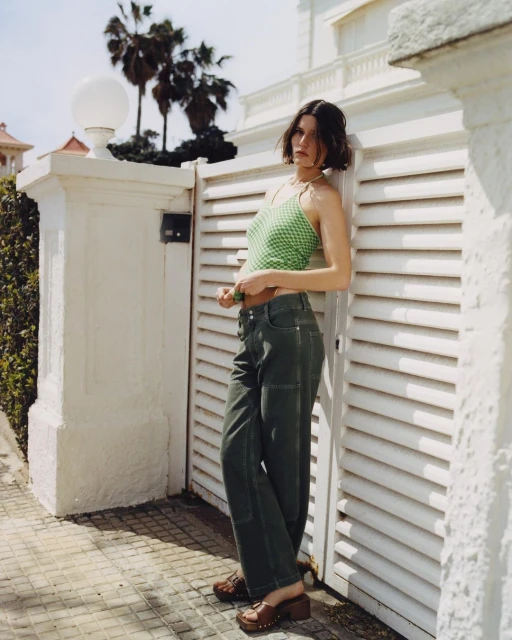 A brunette woman in a green tank top and green pants poses against a white wall with palm trees in the background on a sunny day.