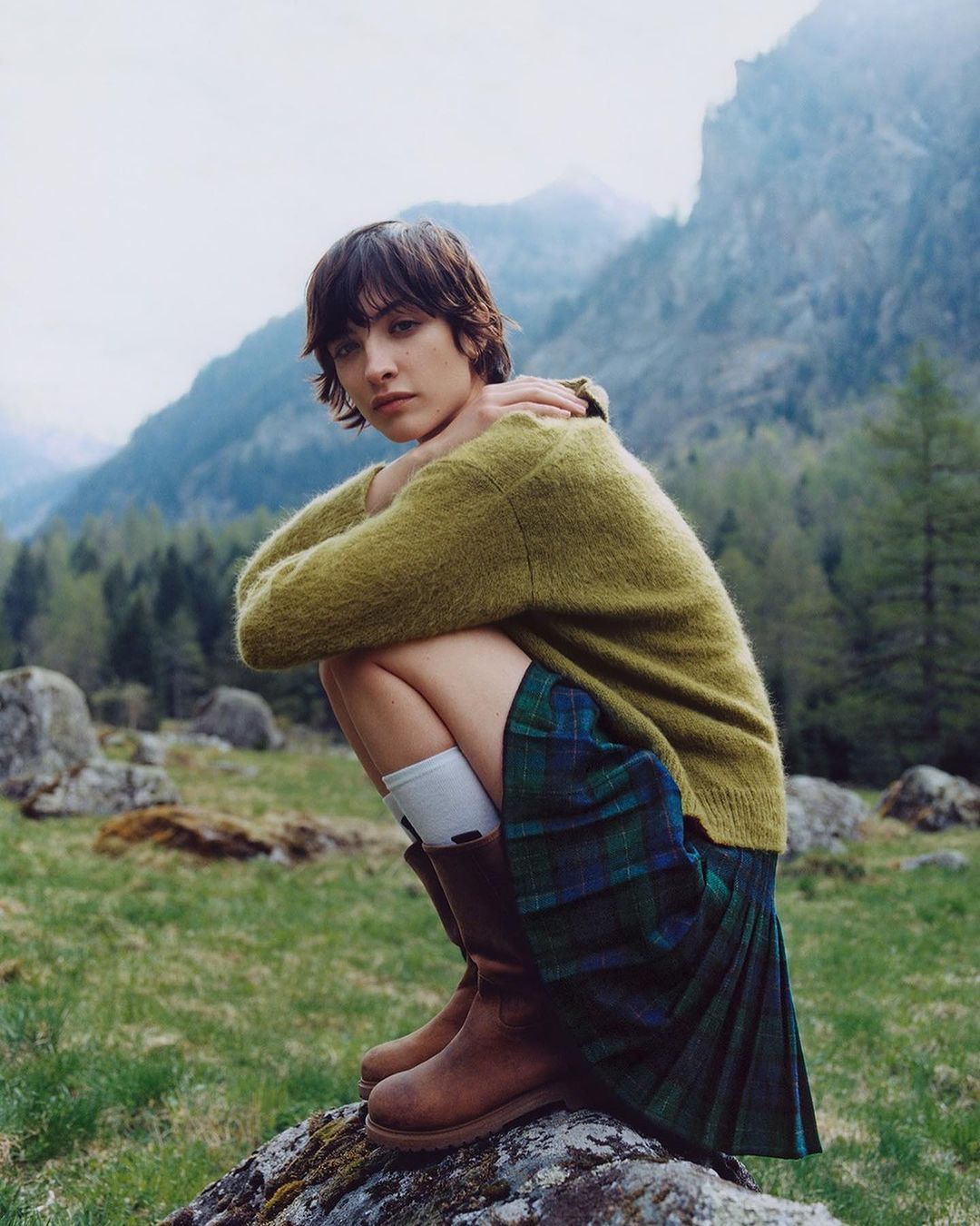 A young adult woman with short dark hair, wearing a green sweater, plaid skirt, and brown boots, poses outdoors in a mountainous setting.