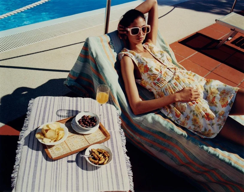 Adult woman relaxing on a striped lounger by a pool with a snack table holding olives, crackers, chips, and a glass of juice.