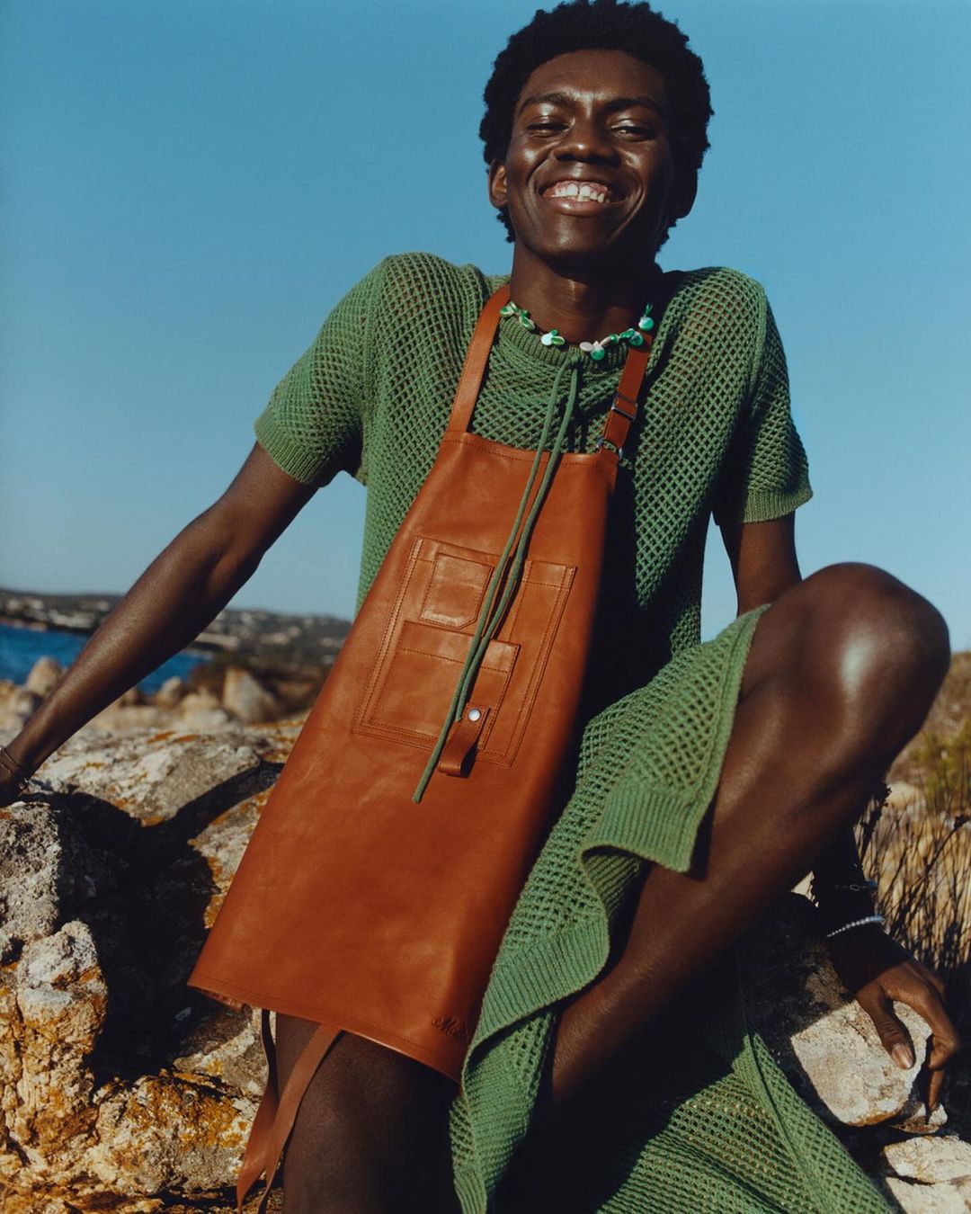 Smiling Black man poses on rocks with a leather apron, green knit outfit, and beaded necklace under a clear blue sky with ocean background.