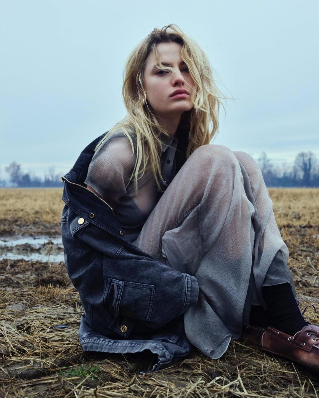 A blonde woman sits in a harvested field wearing a sheer gray dress and denim jacket, gazing at the camera with a neutral expression.